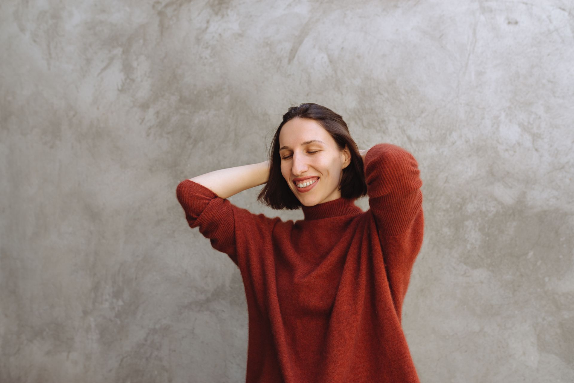 Una donna con un maglione rosso è in piedi davanti a un muro grigio con le mani dietro la testa.