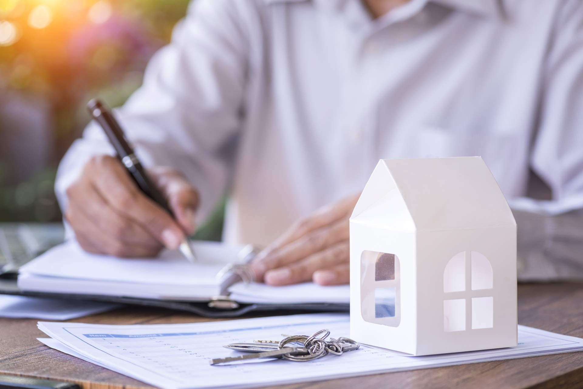 A man is writing on a piece of paper next to a model house and keys.