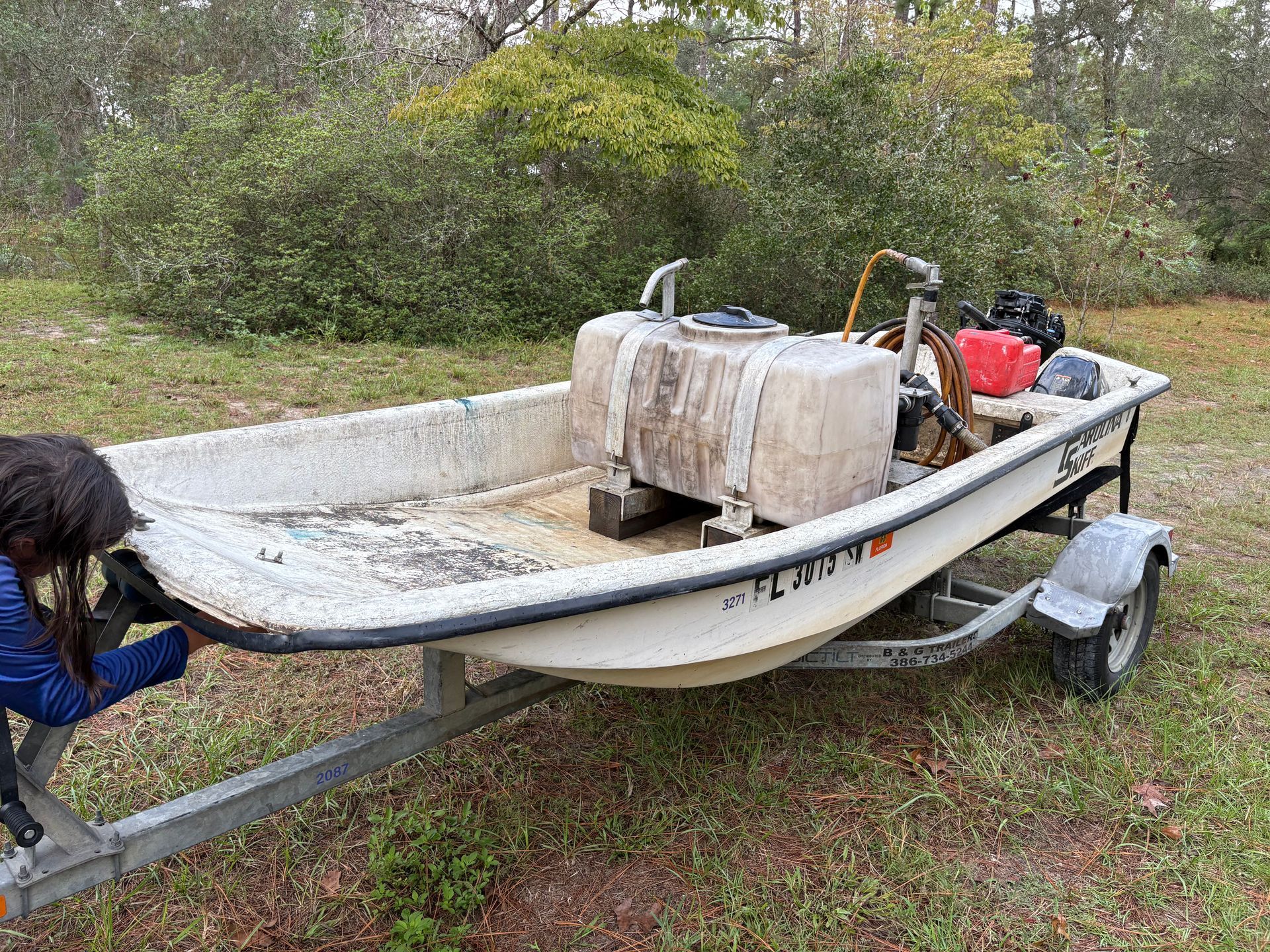 Boat on a trailer, with a person adjusting something near the boat. Outdoors, with foliage in the background.