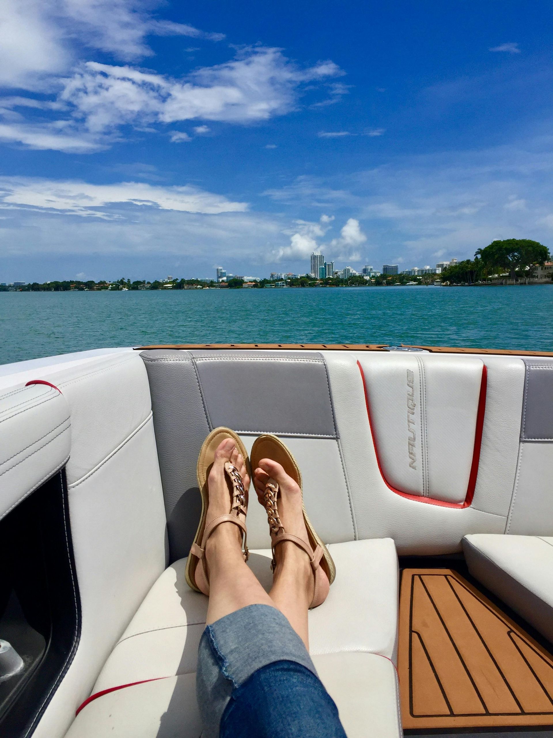 Feet in sandals on a boat, city skyline in the distance, sunny blue sky.