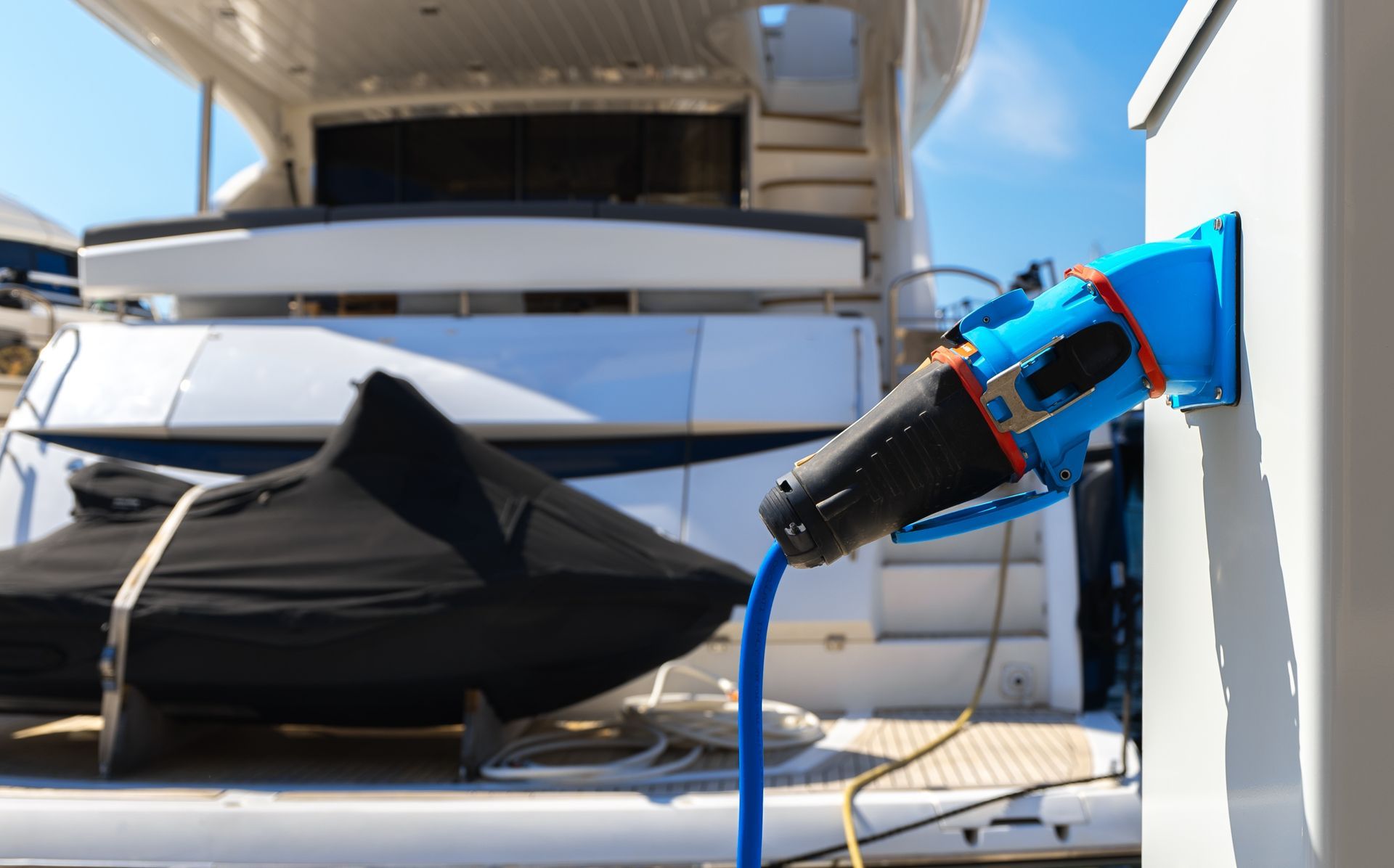 Blue power cord plugged into a white dockside electrical outlet, with a covered boat in the background.