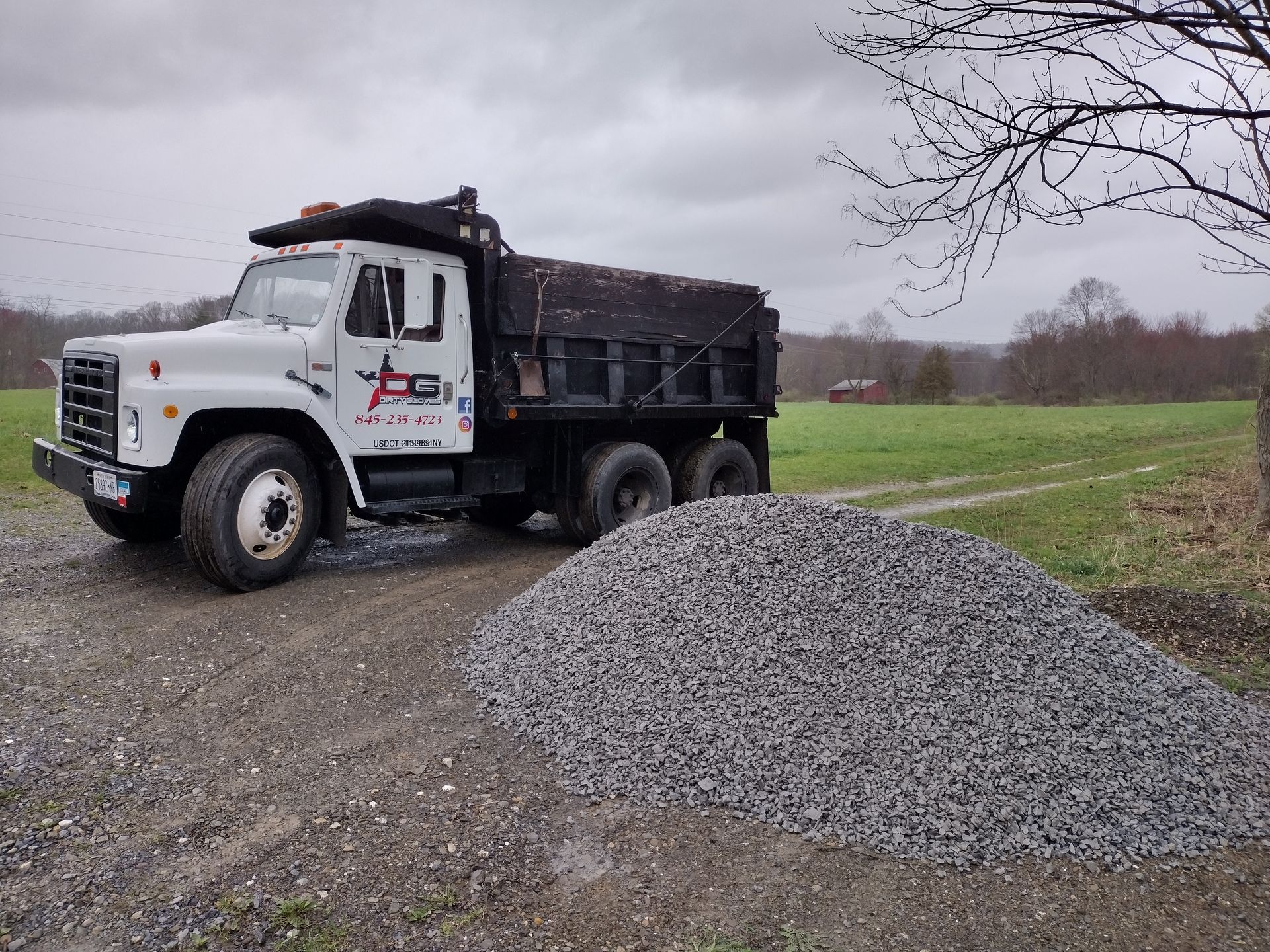 White dump truck beside a pile of gravel on a dirt road, with a field and cloudy sky in the background.