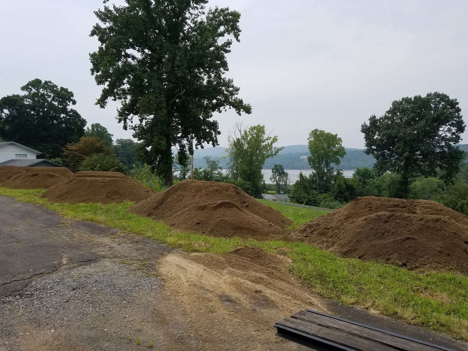 Piles of dirt along a road, trees, and a body of water on a cloudy day.
