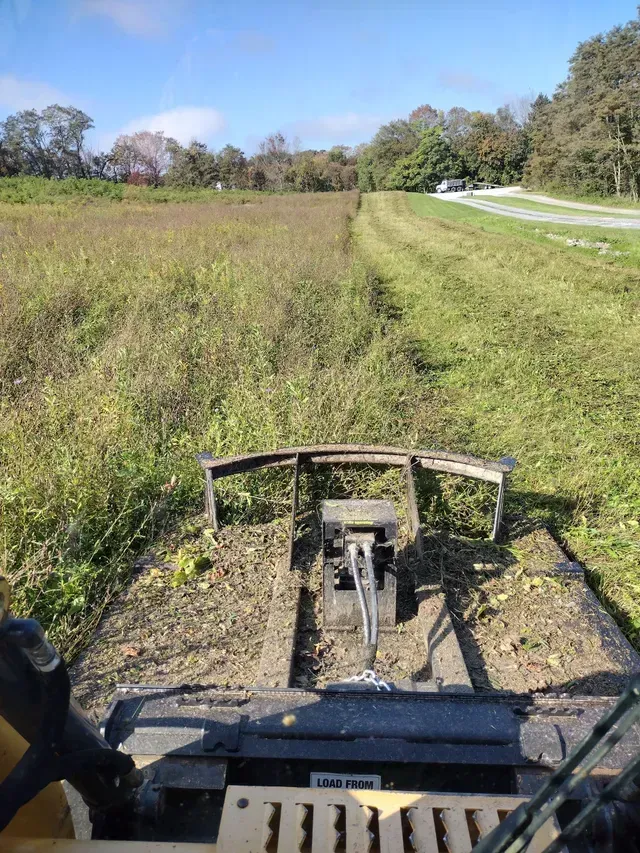A small tractor mowing a field. Green grass is cut in rows. Brown and green weeds remain uncut in the foreground. A small tractor mowing a field. Green grass is cut in rows. Brown and green weeds remain uncut in the foreground.