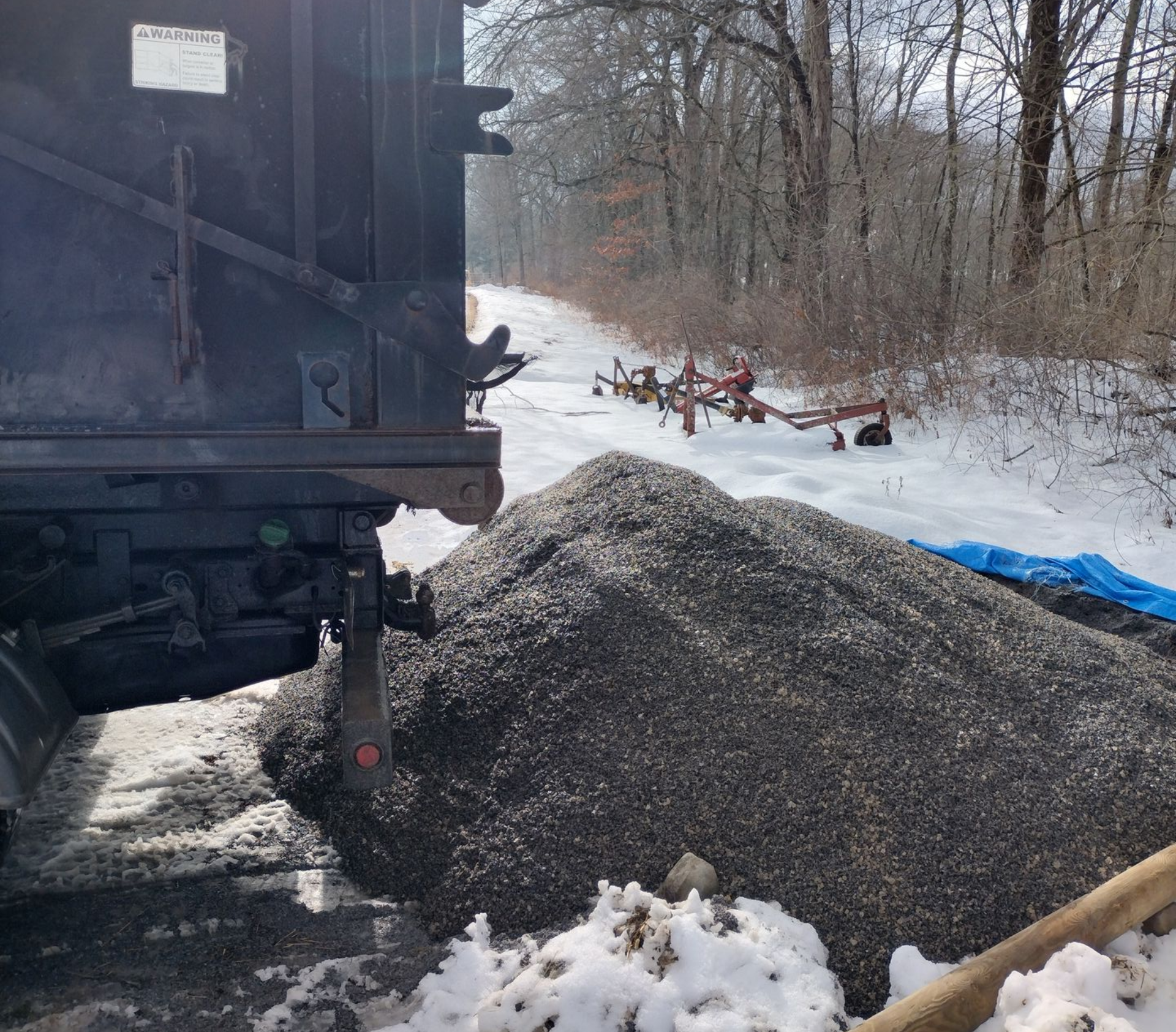 Dump truck unloading gravel on a snow-covered rural road. Farm equipment is visible in the background.