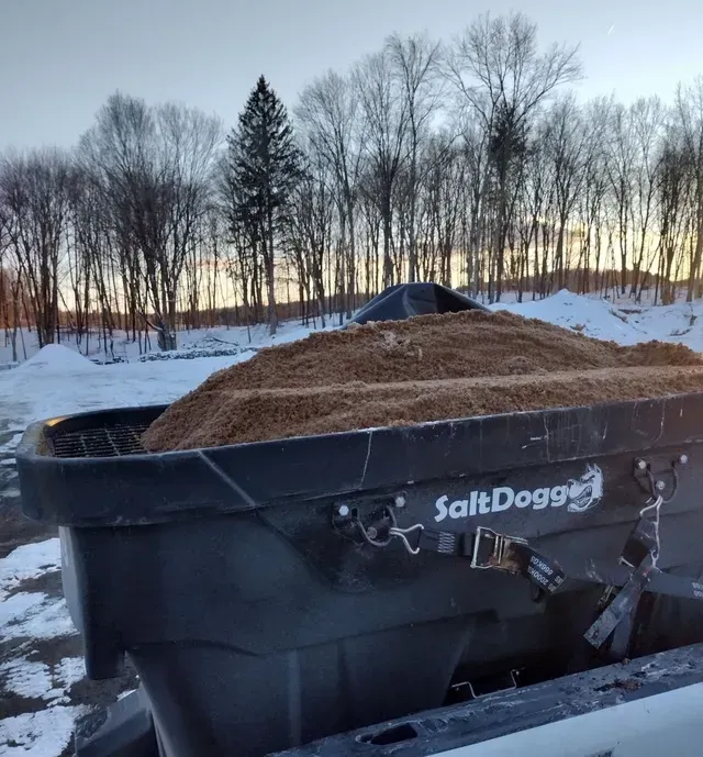 A black SaltDogg salt spreader filled with sand, mounted on a truck bed against a snowy landscape with bare trees. A black SaltDogg salt spreader filled with sand, mounted on a truck bed against a snowy landscape with bare trees.