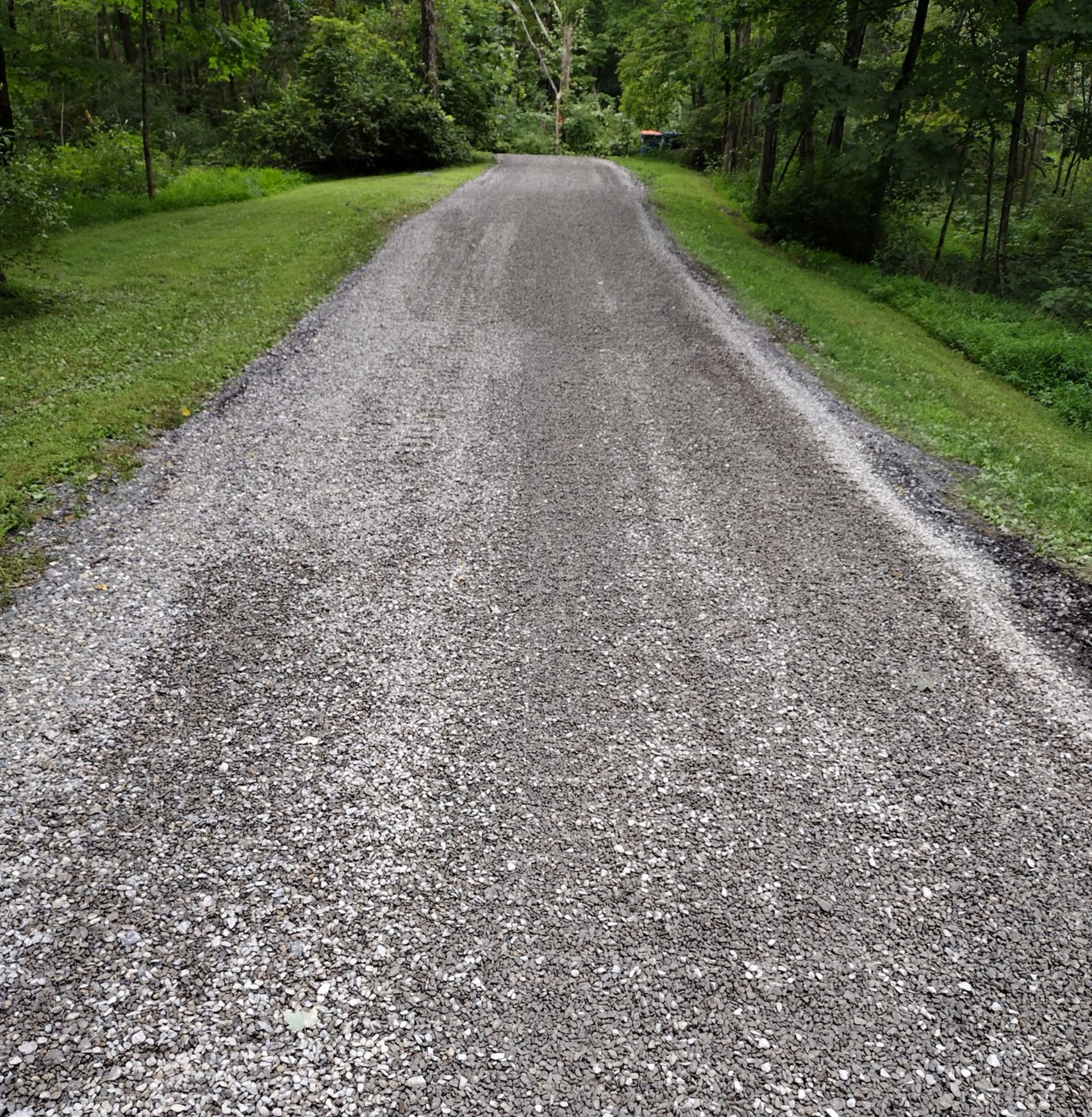 Gravel driveway curves uphill toward a red barn, with a yellow skid steer parked to the side. A straight, gravel driveway leads through a grassy, wooded landscape under an overcast sky.