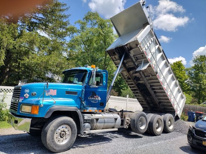 Blue dump truck dumping gravel on a residential street under a sunny sky.