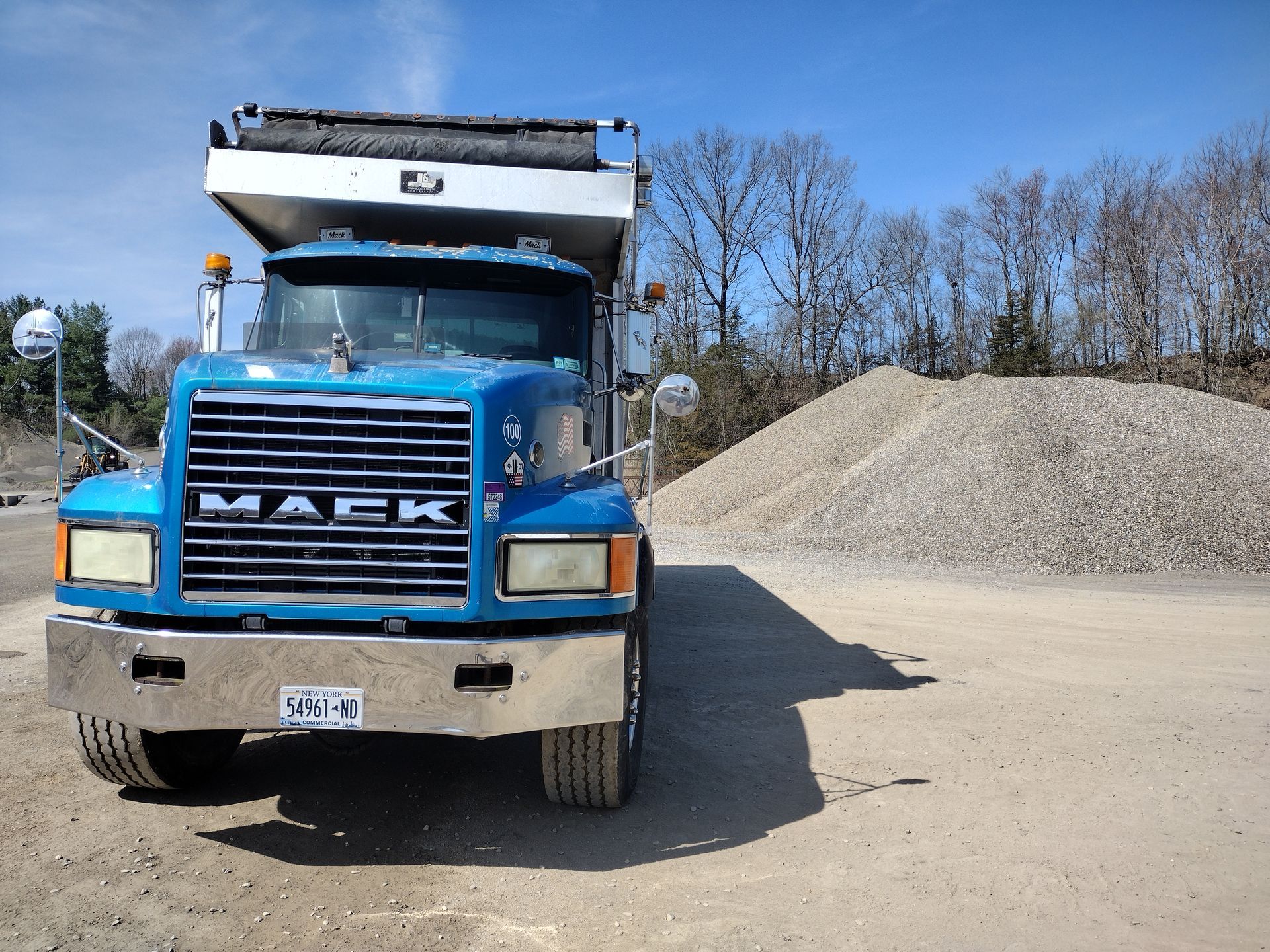 Blue Mack dump truck parked near a gravel pile under a blue sky.
