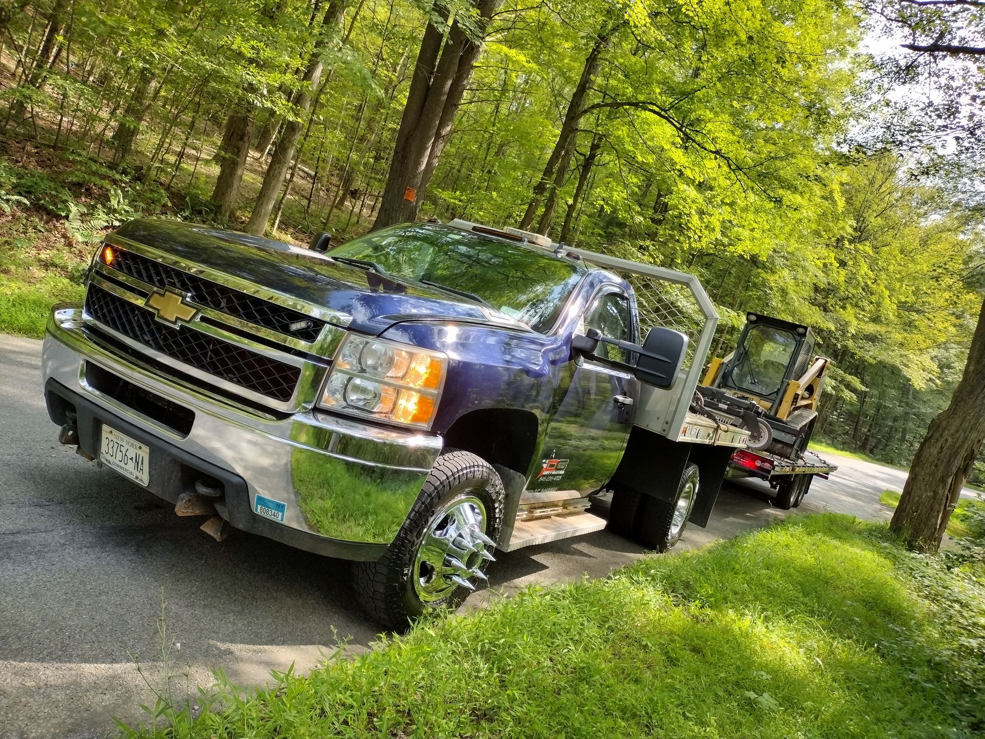 Dark blue Chevy dually truck pulling a trailer on a gravel road, surrounded by trees and grass.