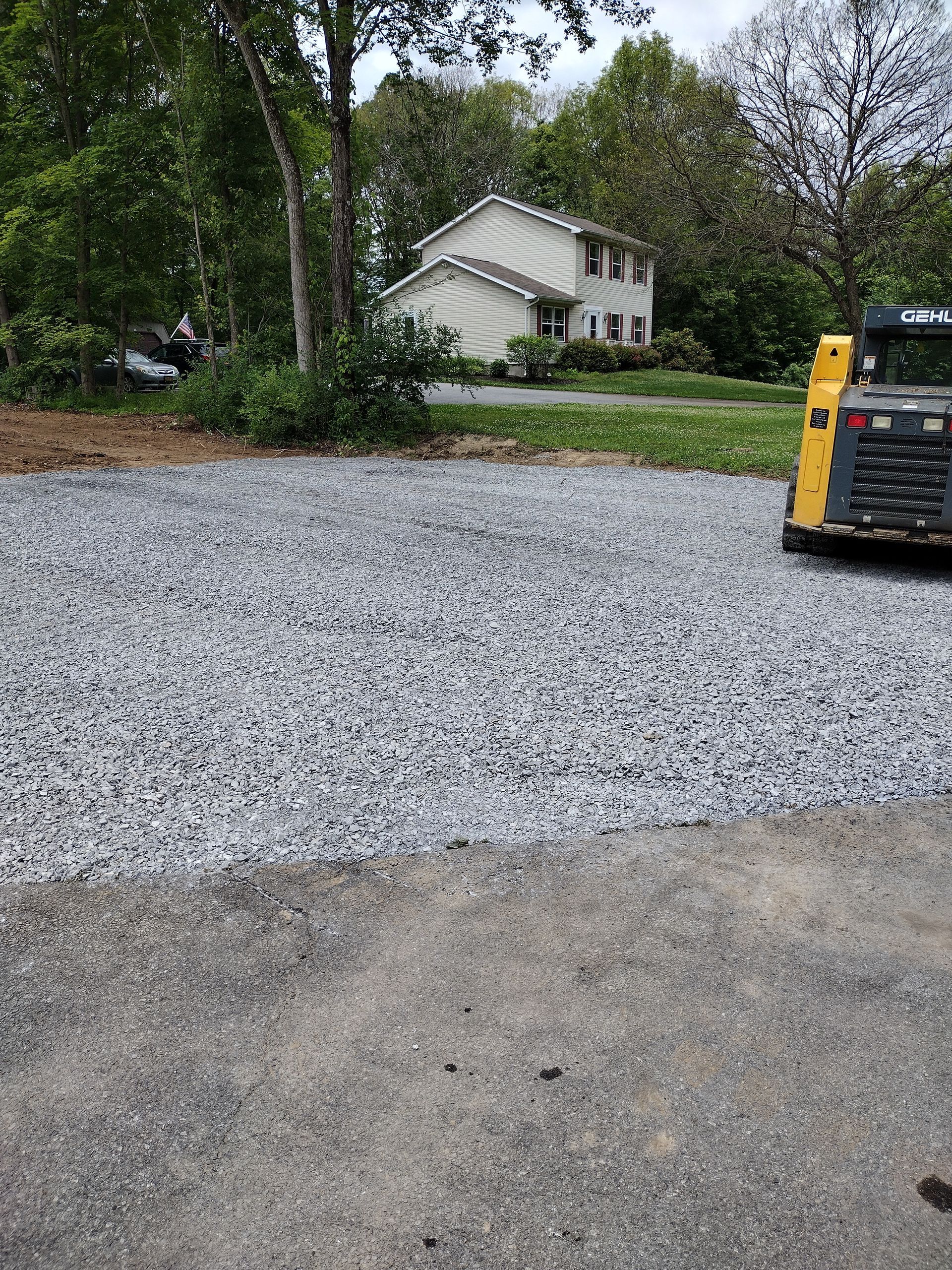 Gravel parking area next to asphalt. A two-story house is in the background. A skid-steer loader is visible.