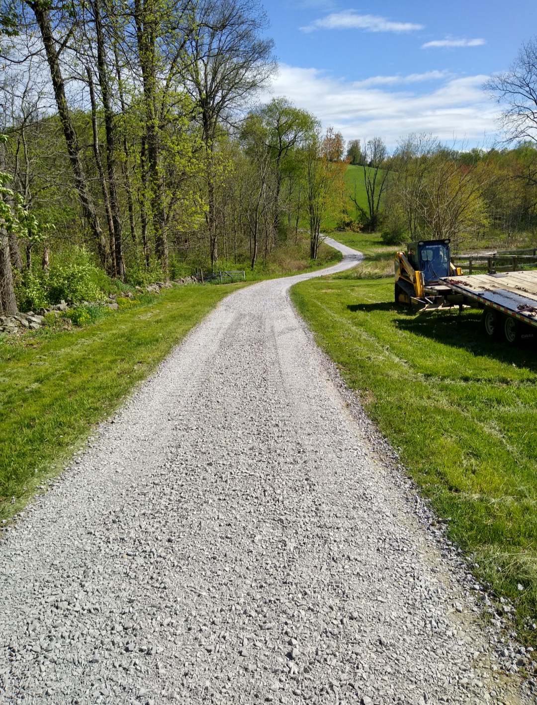 Gravel driveway winding uphill through grassy field, small construction vehicle on right, trees and blue sky.