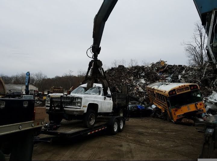 Crane lifting a white pickup truck onto a trailer in a junkyard. Yellow school bus and scrap metal in the background. Crane lifting a white pickup truck onto a trailer in a junkyard. Yellow school bus and scrap metal in the background.