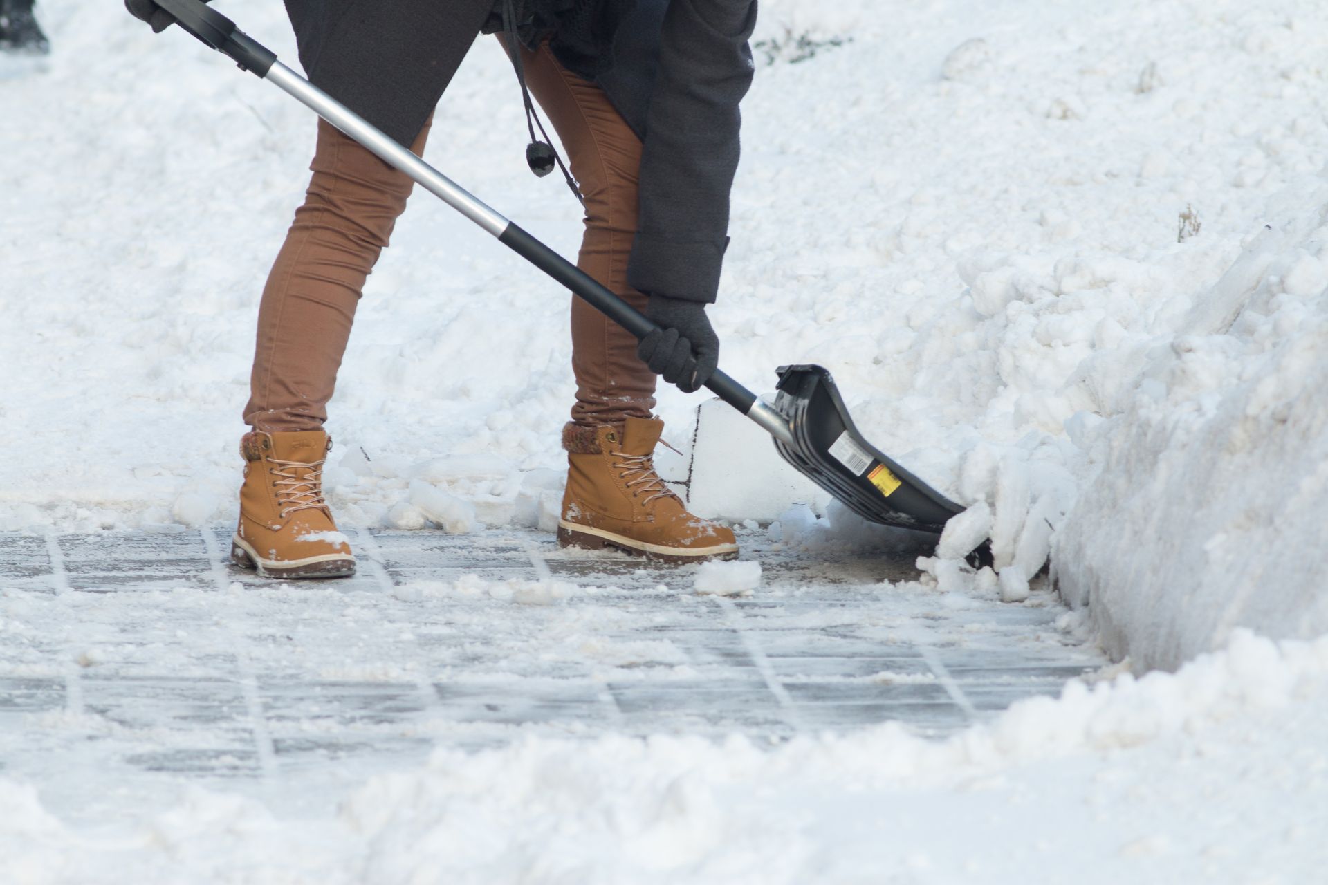 A person in brown pants and tan boots uses a snow shovel to clear snow from a tiled sidewalk. A person in brown pants and tan boots uses a snow shovel to clear snow from a tiled sidewalk.