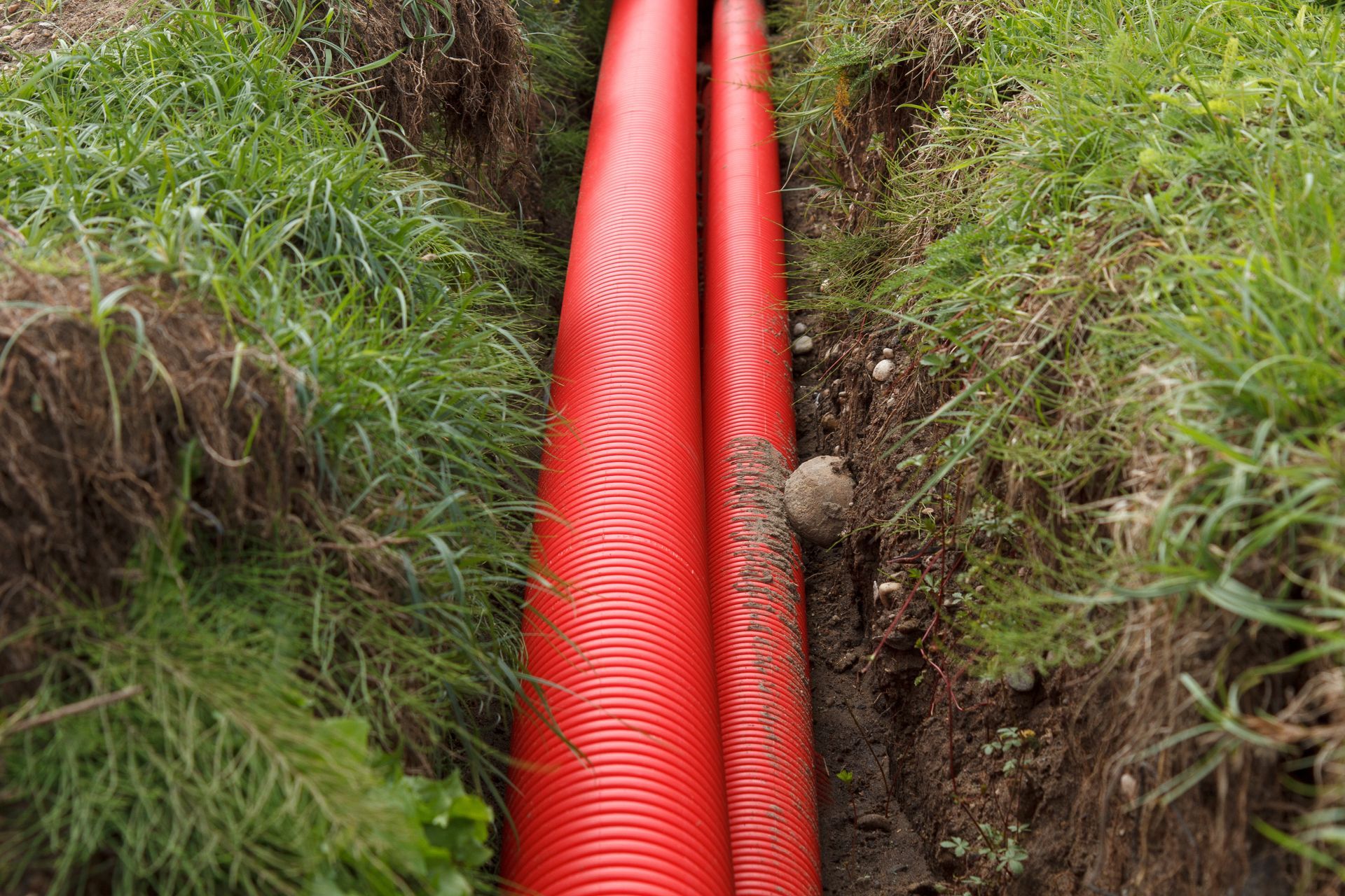 Two red corrugated pipes laid in a narrow trench in grass and dirt. Two red corrugated pipes laid in a narrow trench in grass and dirt.