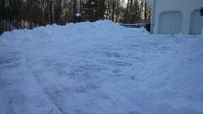 Snow-covered driveway with large snow banks, in front of a white garage on a winter day.