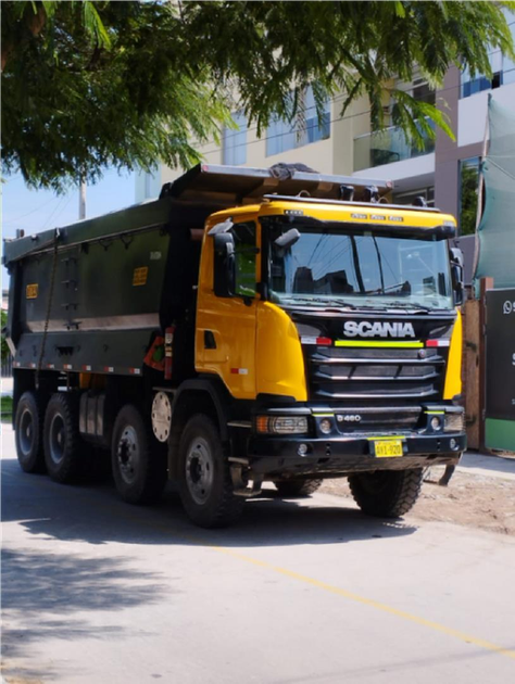 Camión volquete Scania amarillo y negro estacionado en una calle bajo los árboles cerca de edificios.