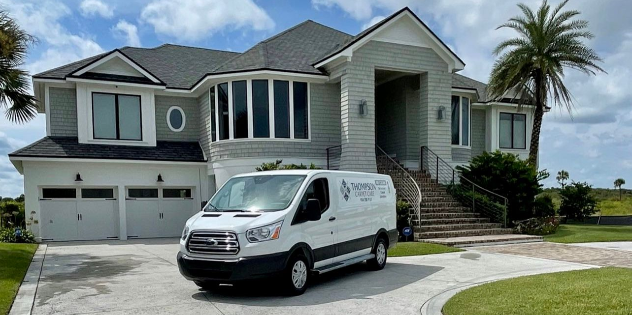 A white service van parked in front of a light gray house. The sky is blue and a palm tree is visible.