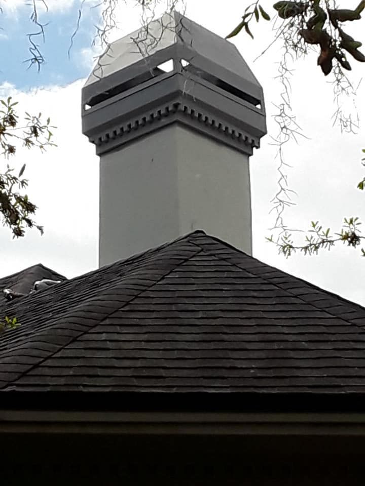 Gray chimney on a dark shingled roof, topped with a geometric cap, against a cloudy sky.