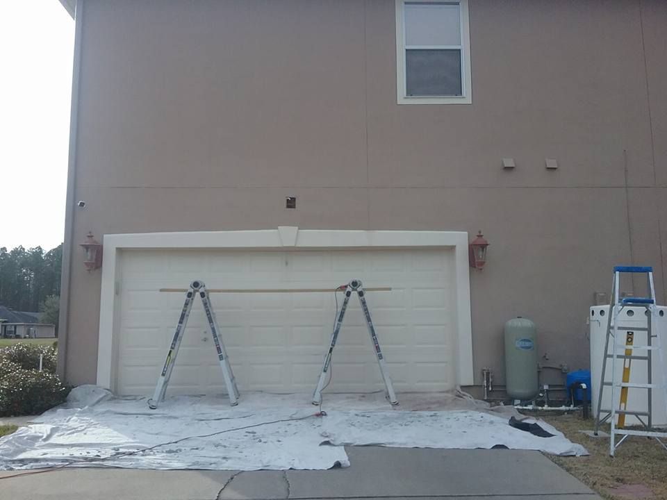 House exterior being painted, beige walls, white trim around garage door, ladders, and protective cloths on the ground.