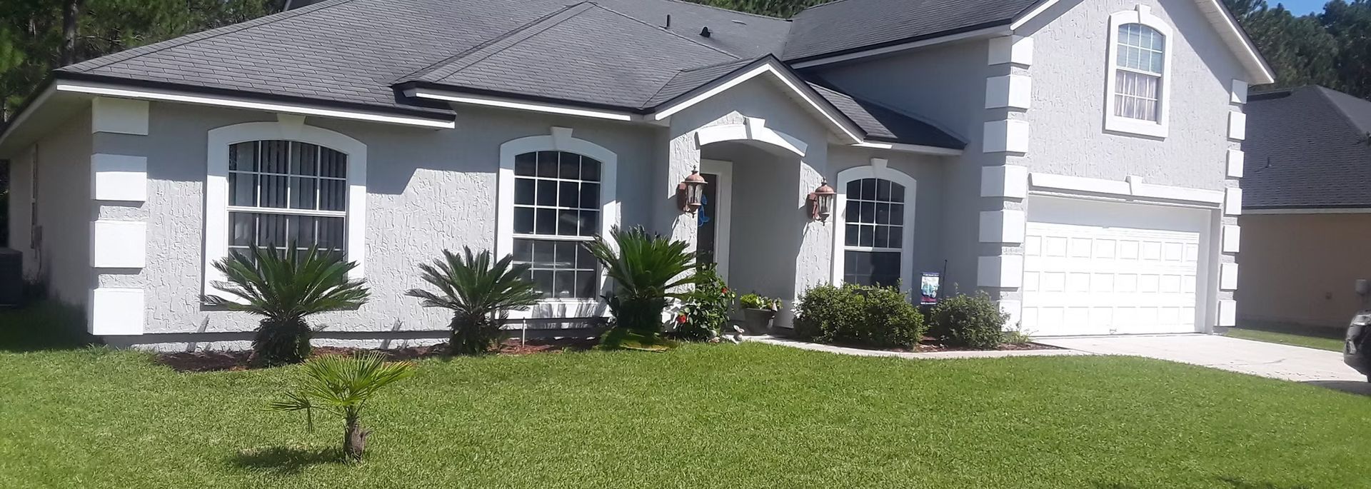 A light gray house with a dark gray roof. The house has a white garage door and is surrounded by green grass and trees.