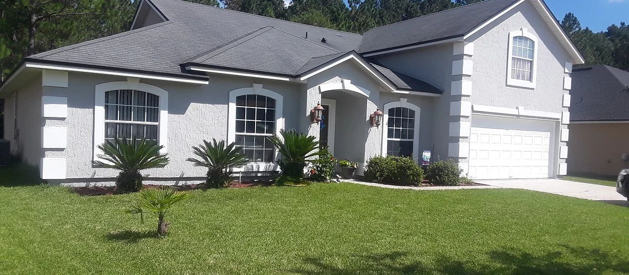 Gray house with white trim, green lawn, trees in the background, blue sky.