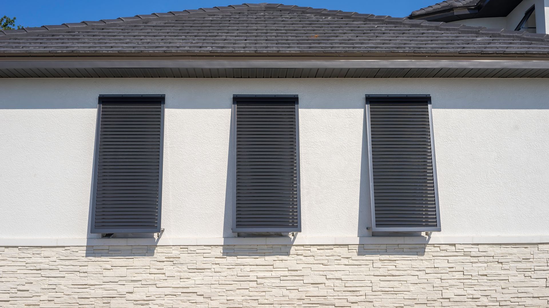 Three windows with dark, slatted shutters on a white stucco wall, below a gray roof.