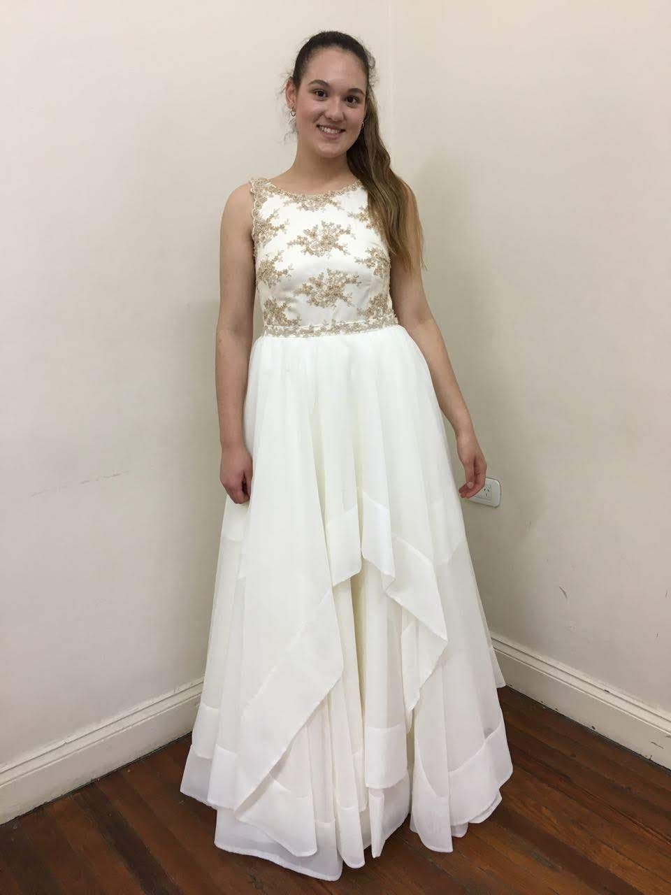 Una mujer con un vestido formal blanco y dorado se encuentra en una habitación con piso de madera.