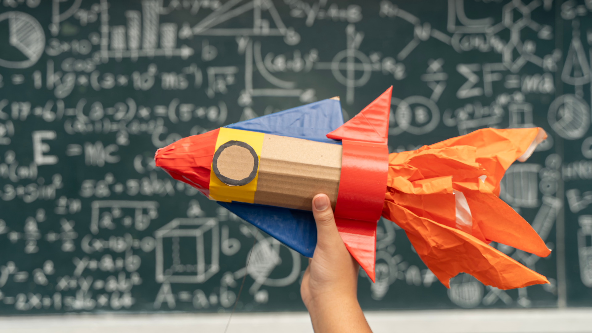 A hand holds a handmade cardboard rocket ship with blue wings, red trim, and orange streamers in front of a chalkboard.