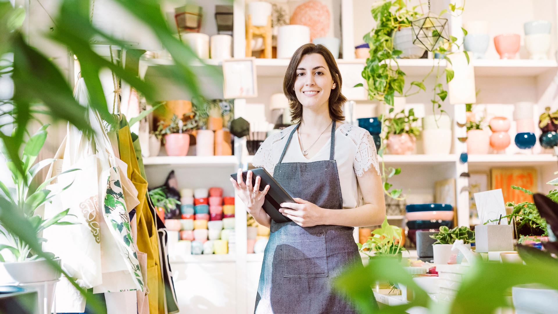 Woman in apron holding a tablet smiles in a plant shop with shelves of plants and pots.