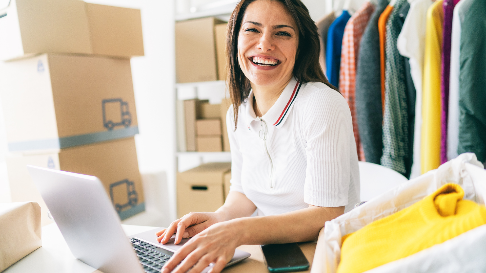 Woman at laptop smiles, surrounded by packages, clothing rack, and packed boxes.
