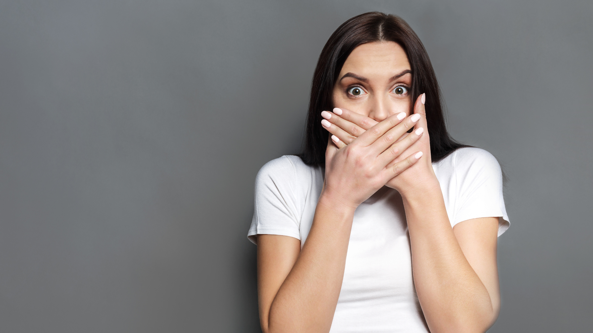 Woman in white shirt, hands covering mouth, wide-eyed, surprised expression, against gray background.