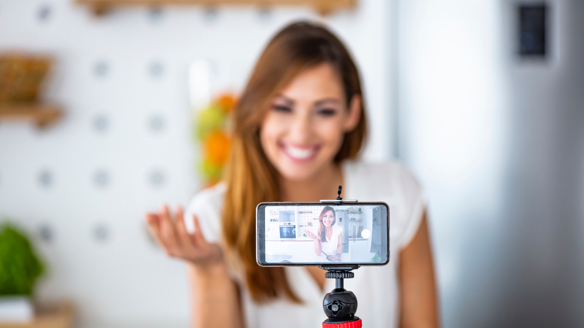 Woman filming with a smartphone on a tripod. She's smiling in a bright kitchen.