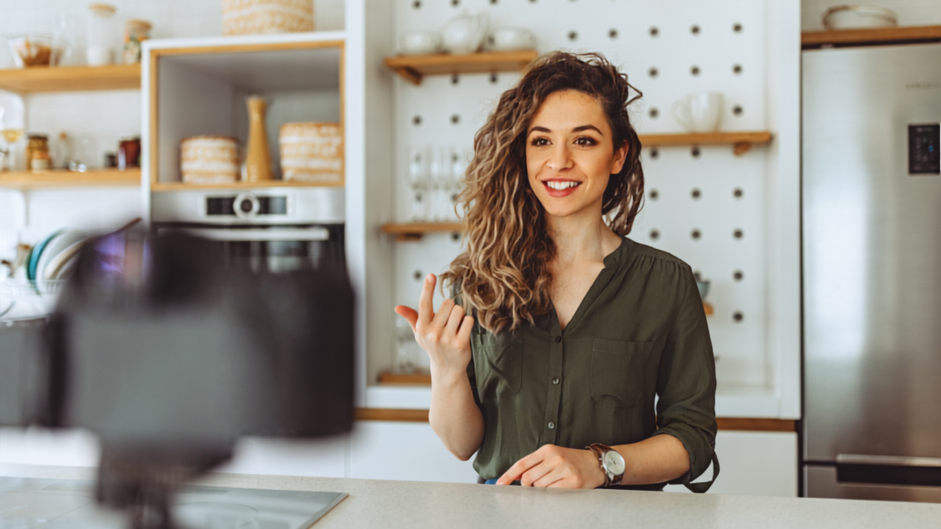Woman smiling and waving in a bright kitchen, with shelves and a blurred camera in the foreground