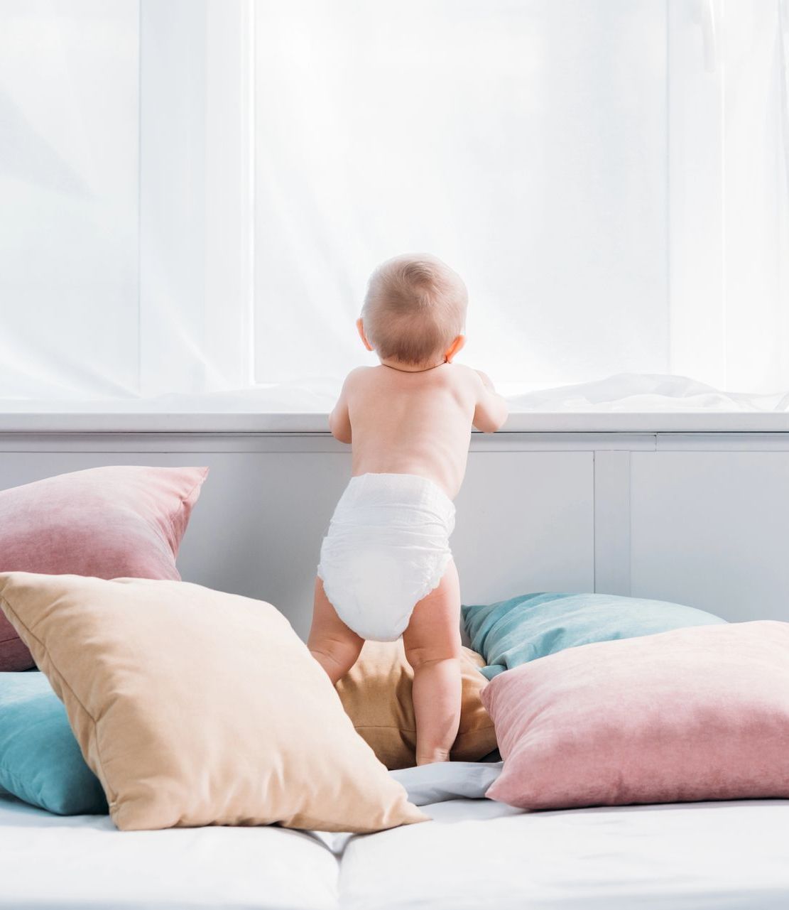 Un bebé con pañal está de pie en una cama mirando por una ventana.
