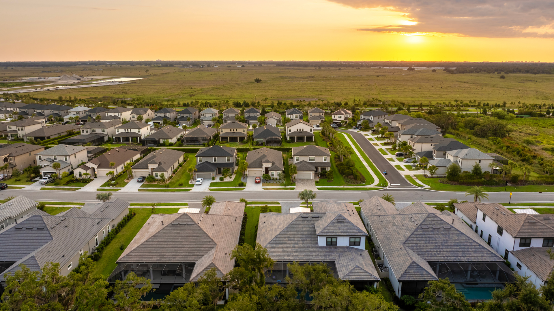 Aerial view of suburban houses at sunset, with a green landscape and road in the background.