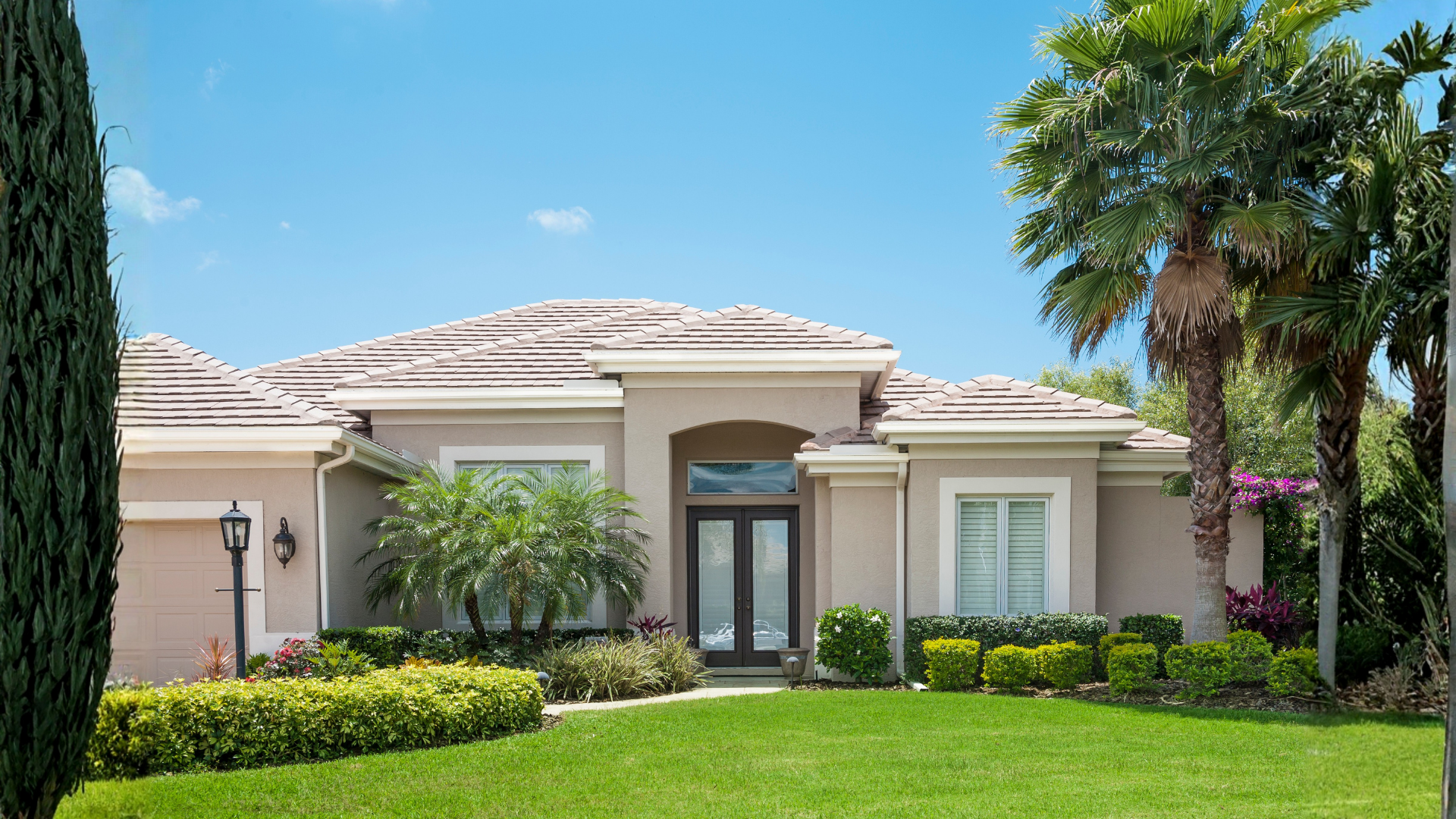 Beige single-story house with green lawn, bushes, palm trees, and a blue sky.