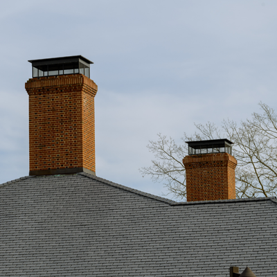 Two brick chimneys with glass caps on a gray shingled roof, against a cloudy sky.