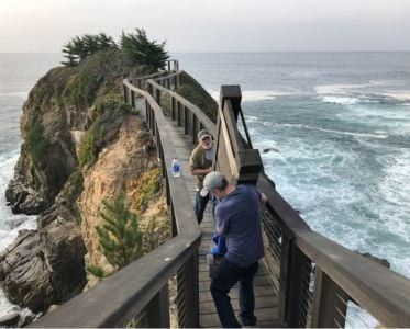 Wooden walkway over ocean, two people walking. Cloudy day, waves crashing on rocks.