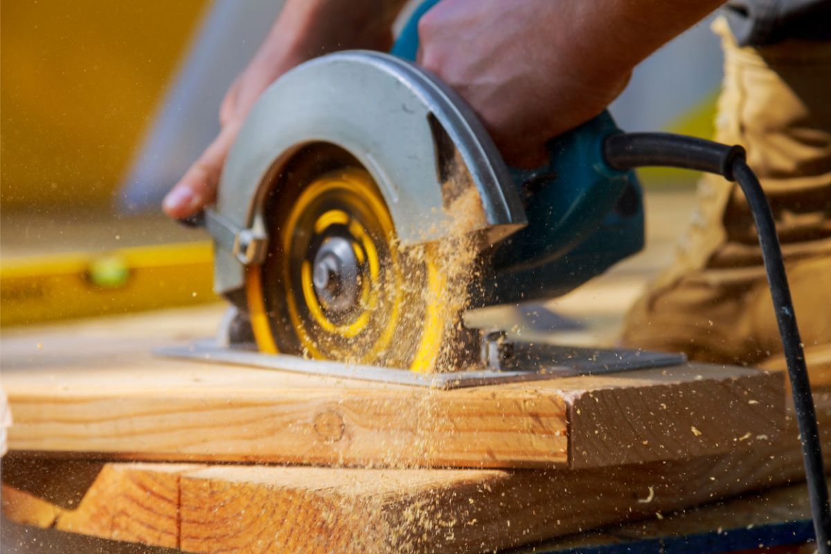 In Rockford, IL, a close-up action shot captures a circular saw as it cuts through a piece of lumber. The hand of a worker steadies the tool, with fine sawdust particles being ejected from the cutting site, creating a dynamic scene filled with movement. The blade, partly obscured by the sawdust cloud, spins rapidly, its teeth biting into the wood's grain. The background shows a blurred yellow level, hinting at precision work in progress. This snapshot highlights the craftsmanship involved in deck building.