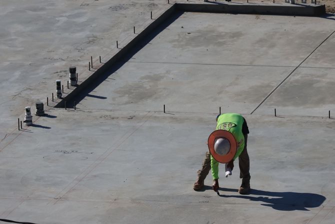Worker in a green vest bending over on a concrete rooftop, handling a red ring near a dark parapet.