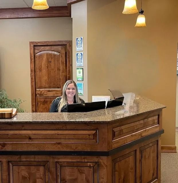 A person smiling behind a wooden reception desk in an office.