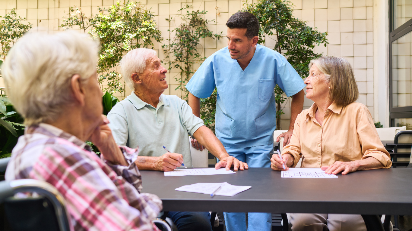 Four older adults with caregiver at table, possibly playing a game. Outdoor setting with greenery.