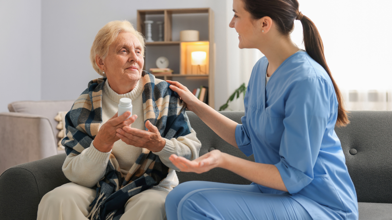 Caregiver assisting an older person with medication in a living room; smiling.