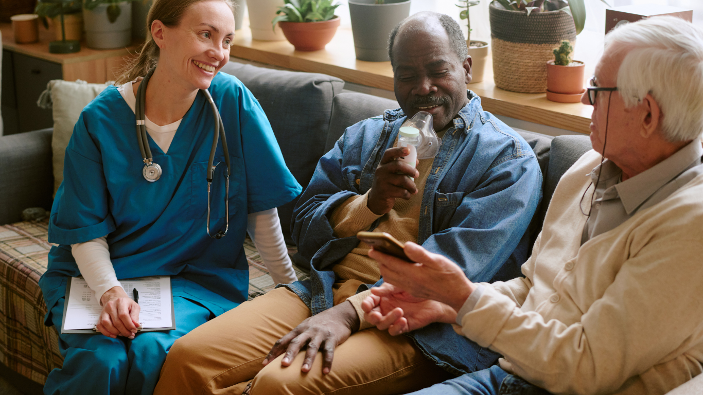 Nurse with stethoscope smiling at man using inhaler, talking with another man on a sofa.