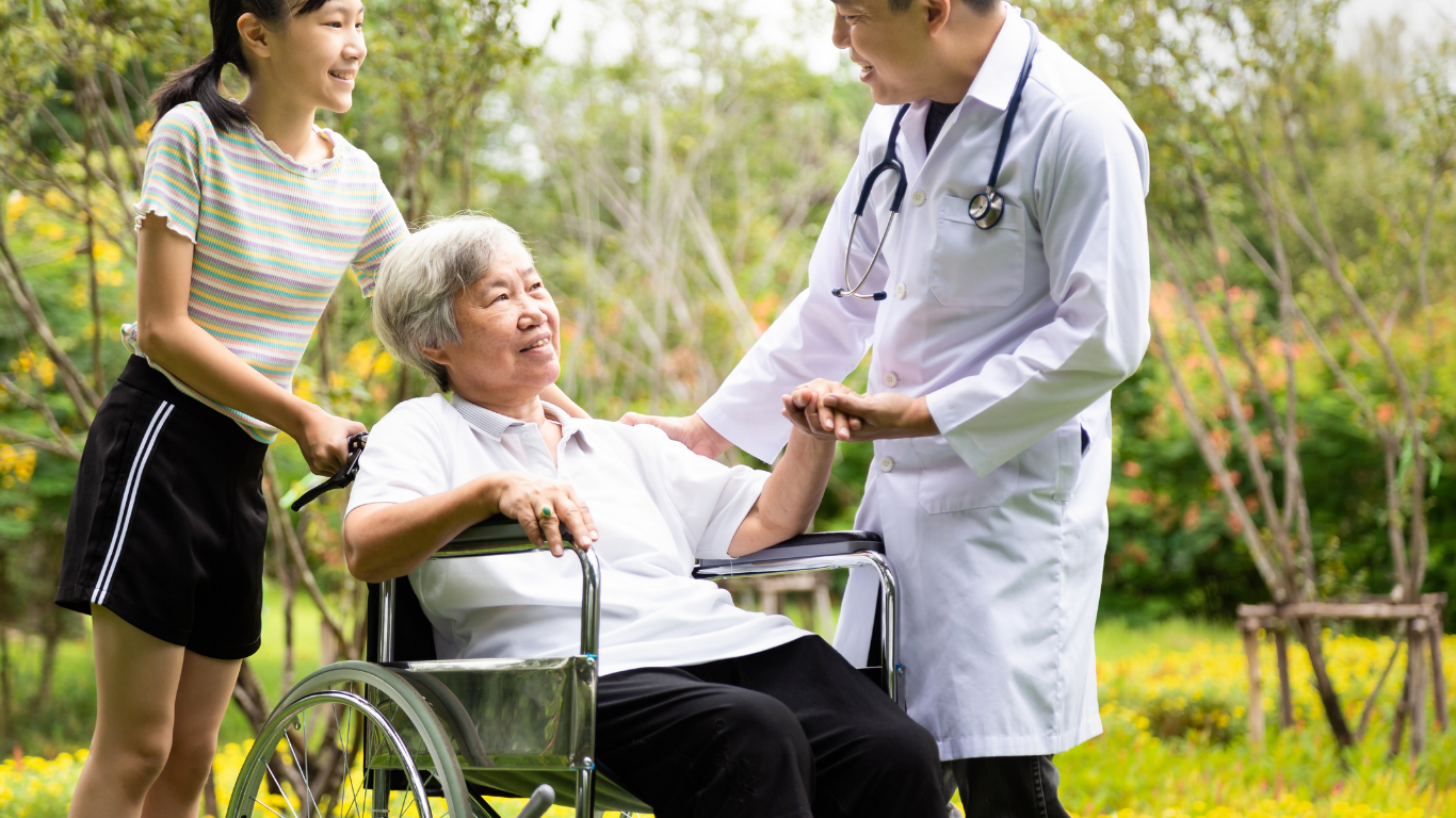 Doctor and young woman assist older person in wheelchair outdoors.