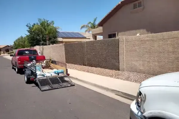 Loaded trailer parked on a residential street beside a red car and beige wall.