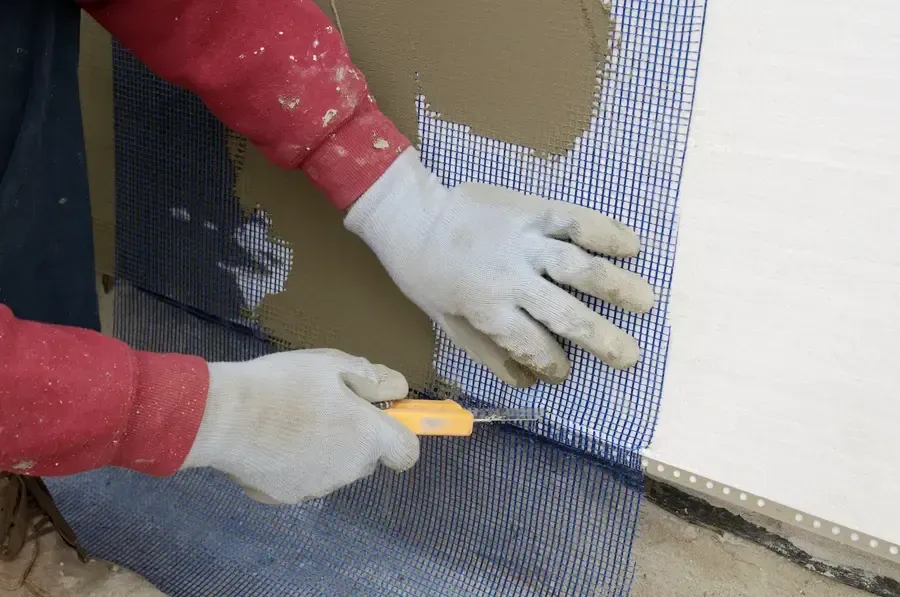 Hands in gloves cutting and fitting black mesh over a wall with a utility knife.