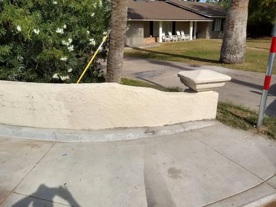 Curved concrete sidewalk beside a cream stucco wall and street, with trees and a house in the background