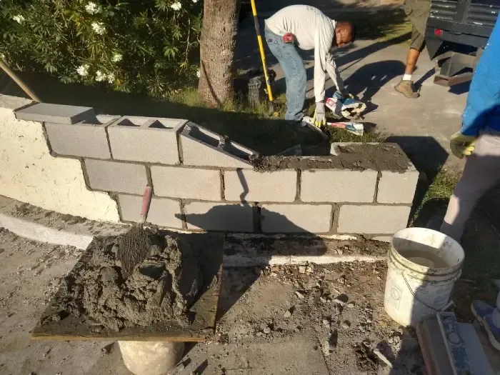 Workers rebuilding a damaged cinder block wall beside a sidewalk, with tools and a bucket nearby.
